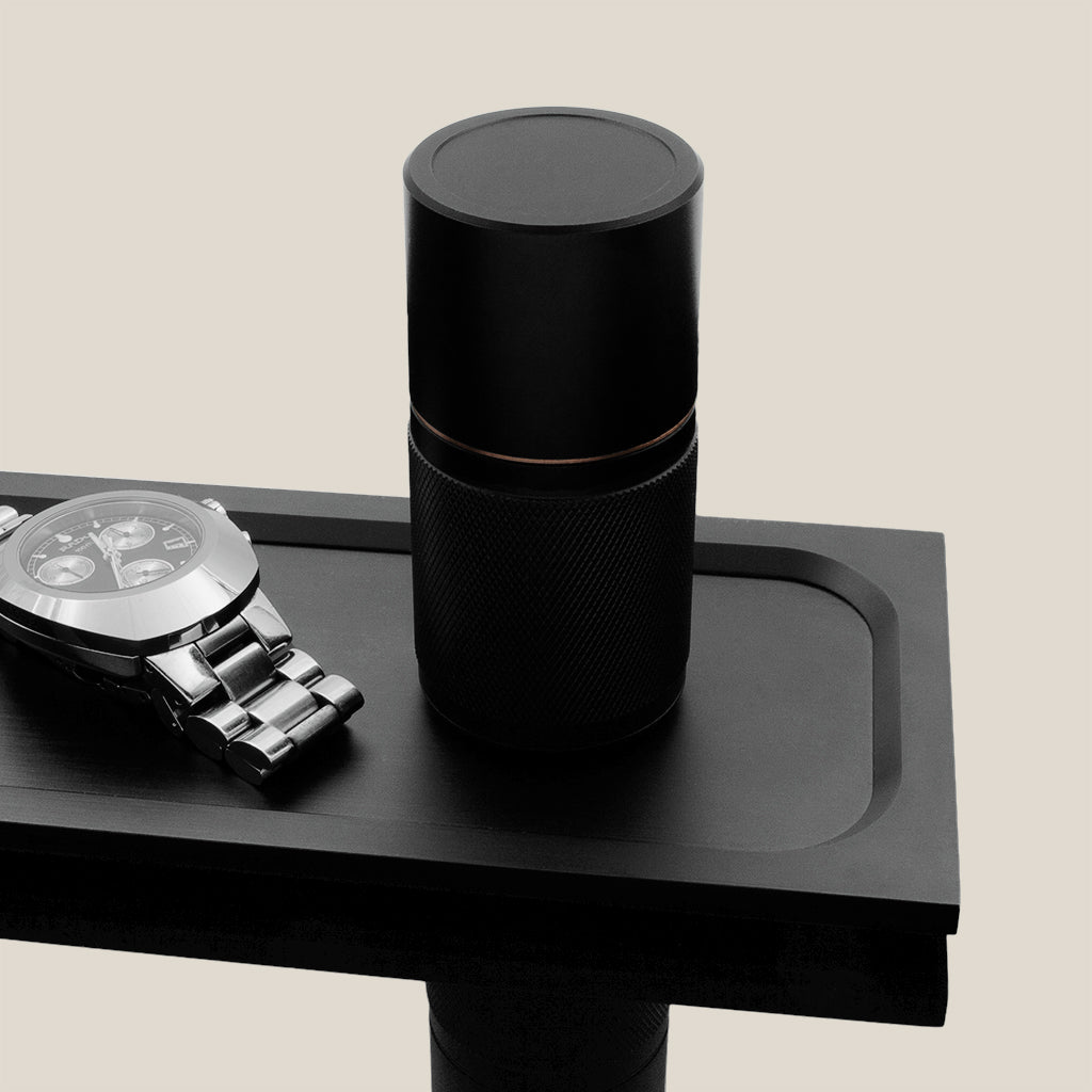 Refillable deodorant with knurled texture and bronze details, standing on a dark plate. Beside a metal watch from the brand Rado.