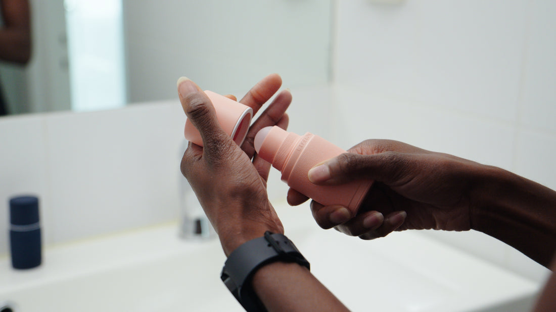 A person’s hand reaching in from the side holding a pink cylindrical deodorant stick against a plain neutral background.
